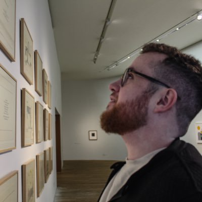 Robert James Gabriel, with a beard and glasses, looking at framed artwork on the wall in a gallery.